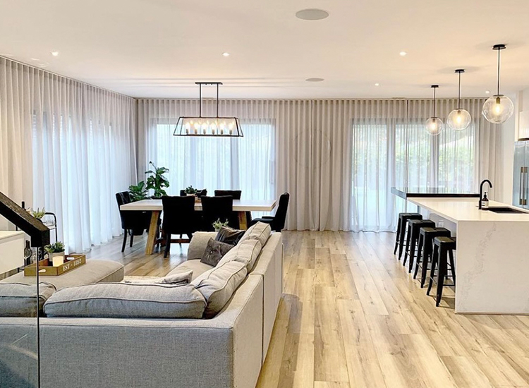 Modern open-plan living space with kitchen and light grey sheer curtains on the corner windows. 