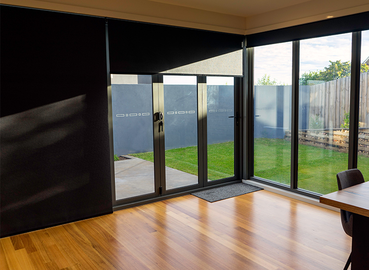 An open plan living room with black linked roller blinds on the bi-fold doors and windows leading to an open backyard.