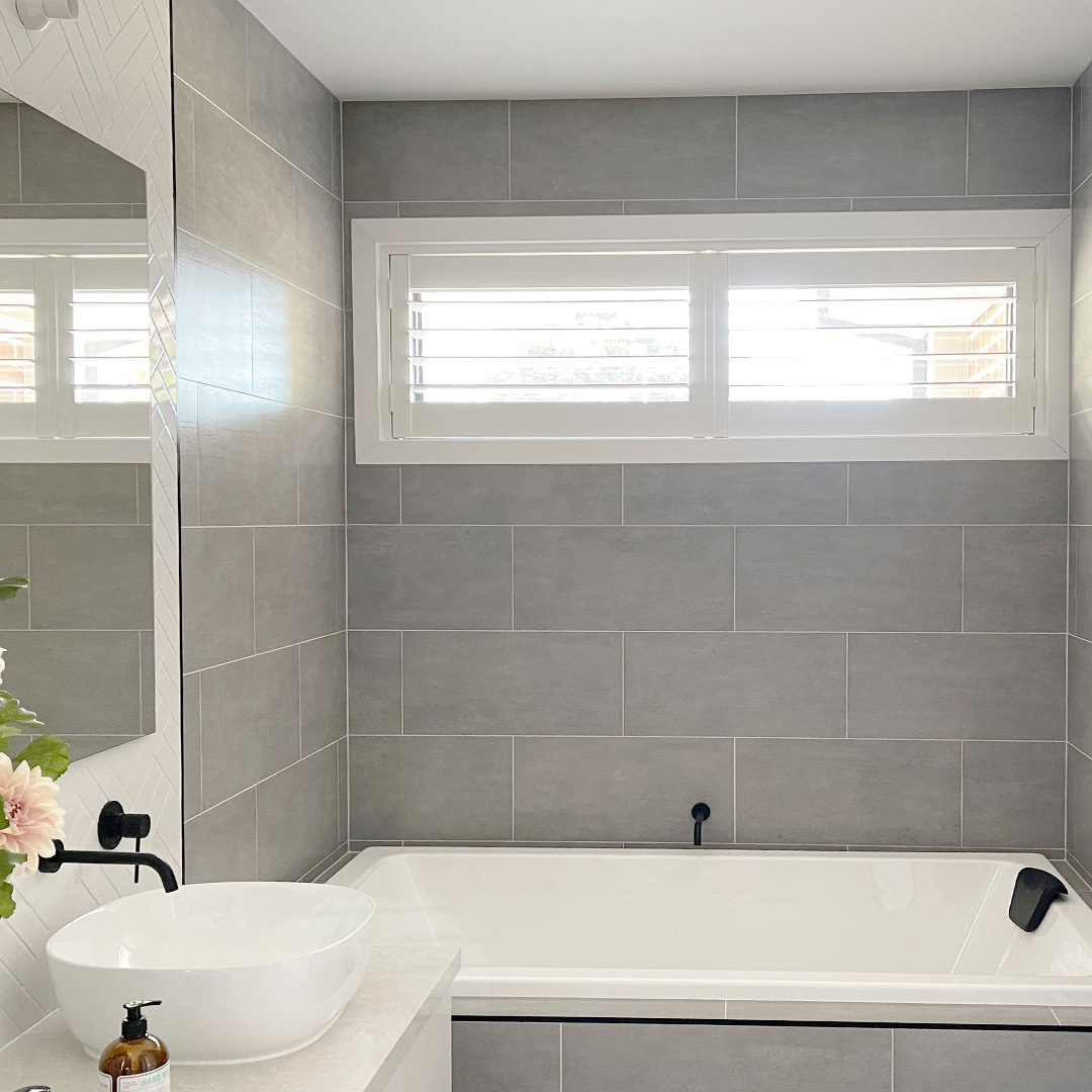 Modern bathroom with grey wall tiles and a white plantation shutter on the highlight window above the bath.